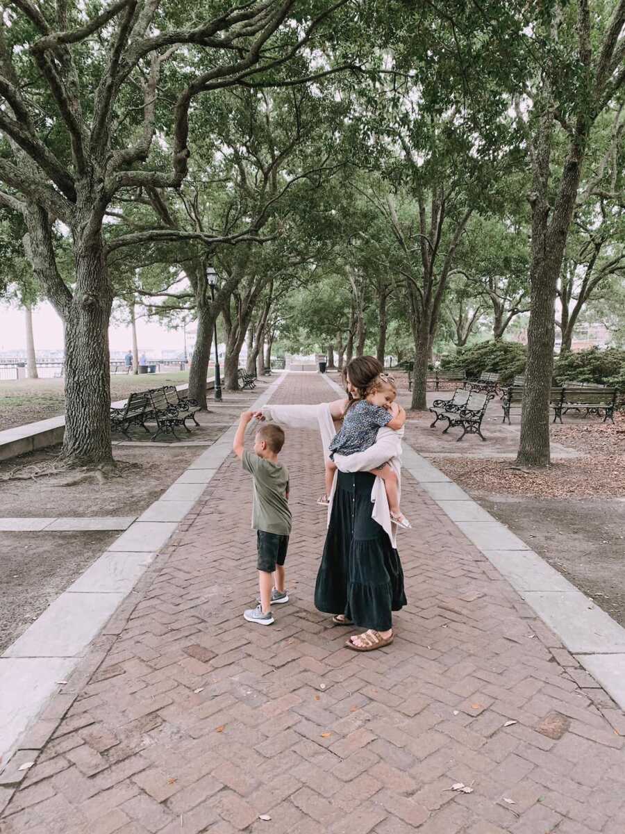 Mom holds daughter and son's hand while standing on pathway in park.