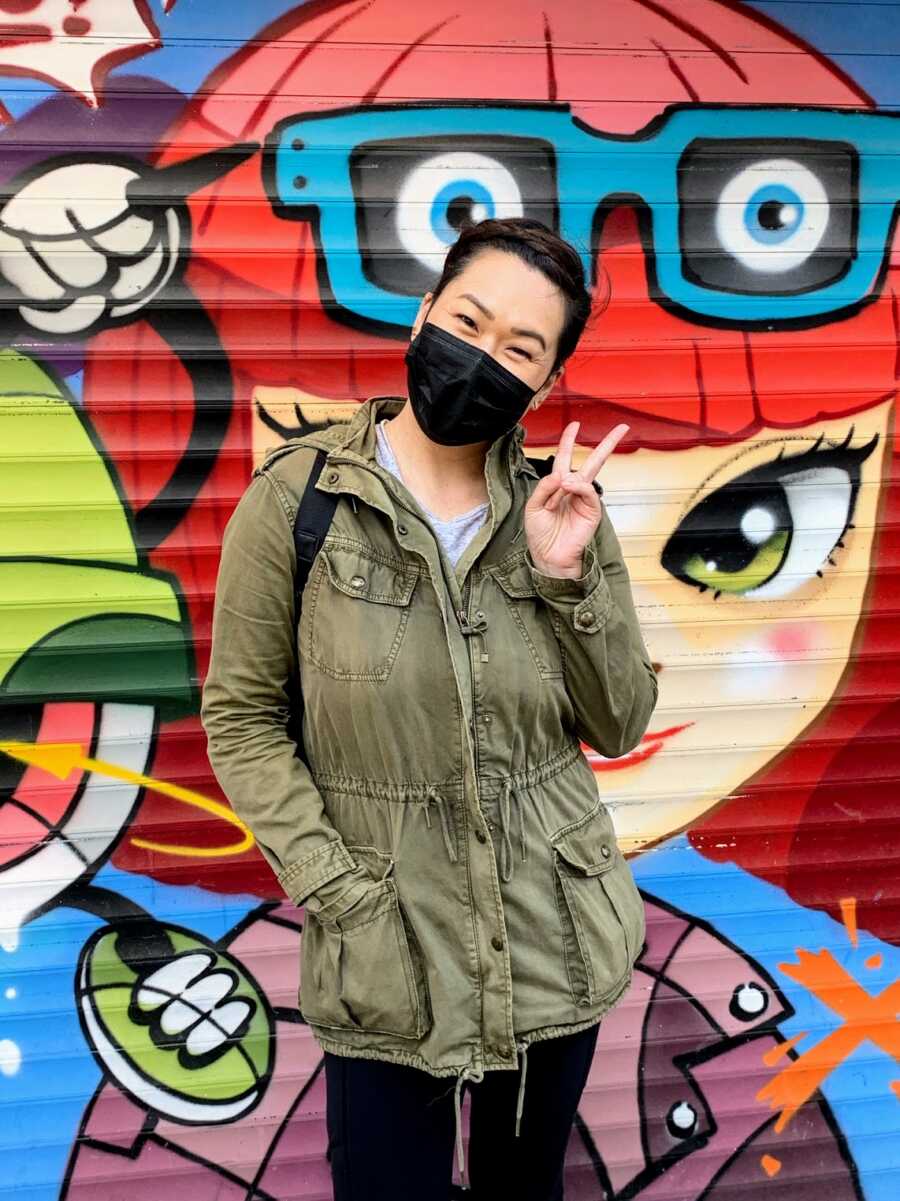 Woman wearing green jacket and black mask stands in front of colorful graffiti mural and makes the peace sign.