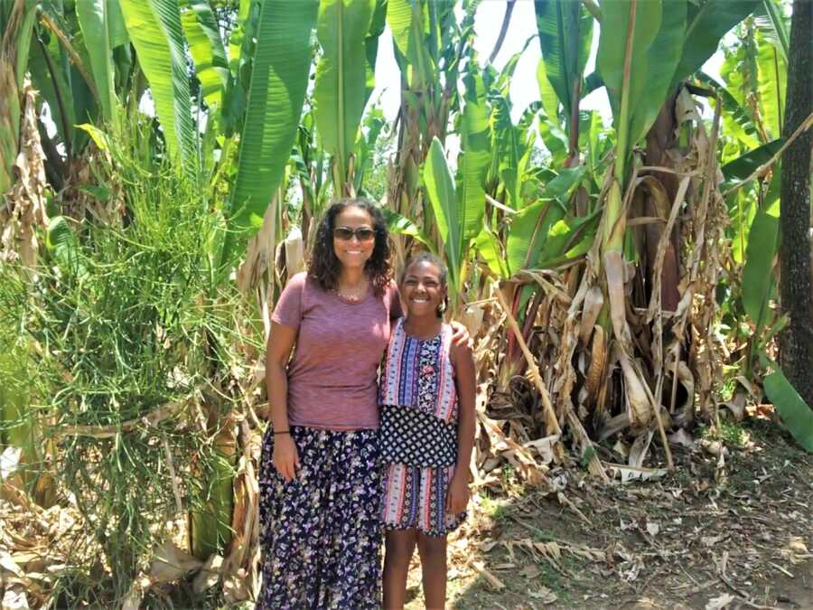 Adoptive mother standing next to her daughter with tall palms in the back