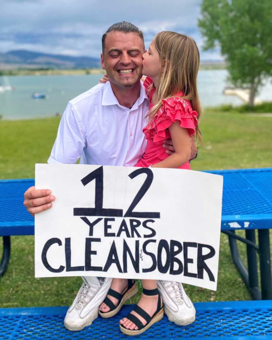 Recovered drug addict sitting with his daughter and holding a sign saying '12 Years Clean and Sober'