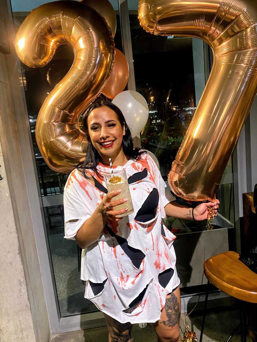 Aboriginal woman in halloween outfit holding a drink and balloons spelling 27