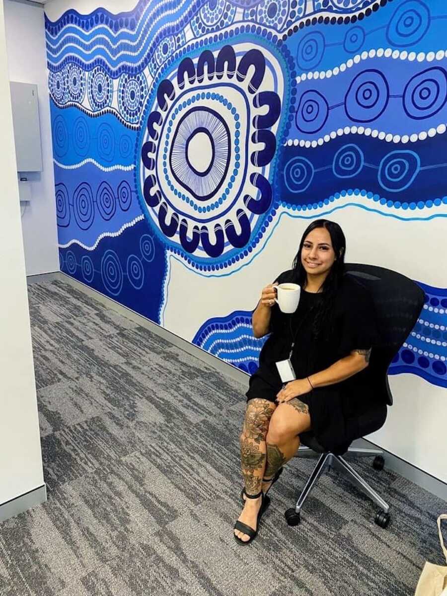 Aboriginal woman sitting in front of art wall with a cup of coffee