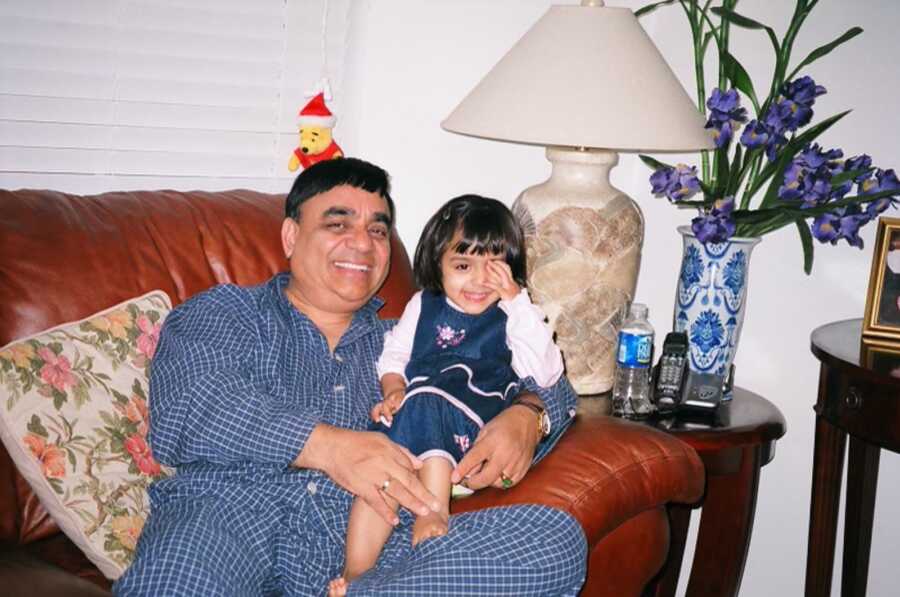 Small Indian girl sits on her father's lap on the couch. 