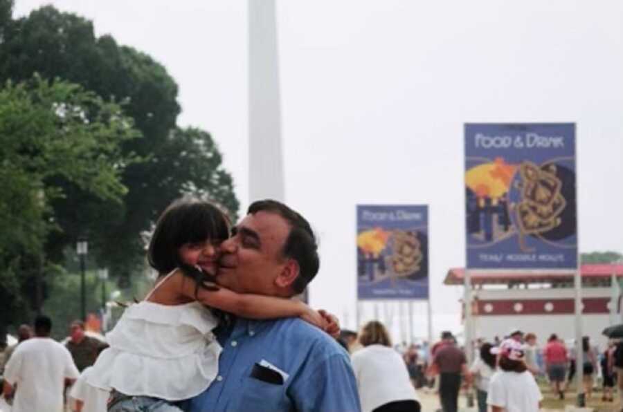 Young girl wraps her arms around her father's neck as he holds her on a busy street.
