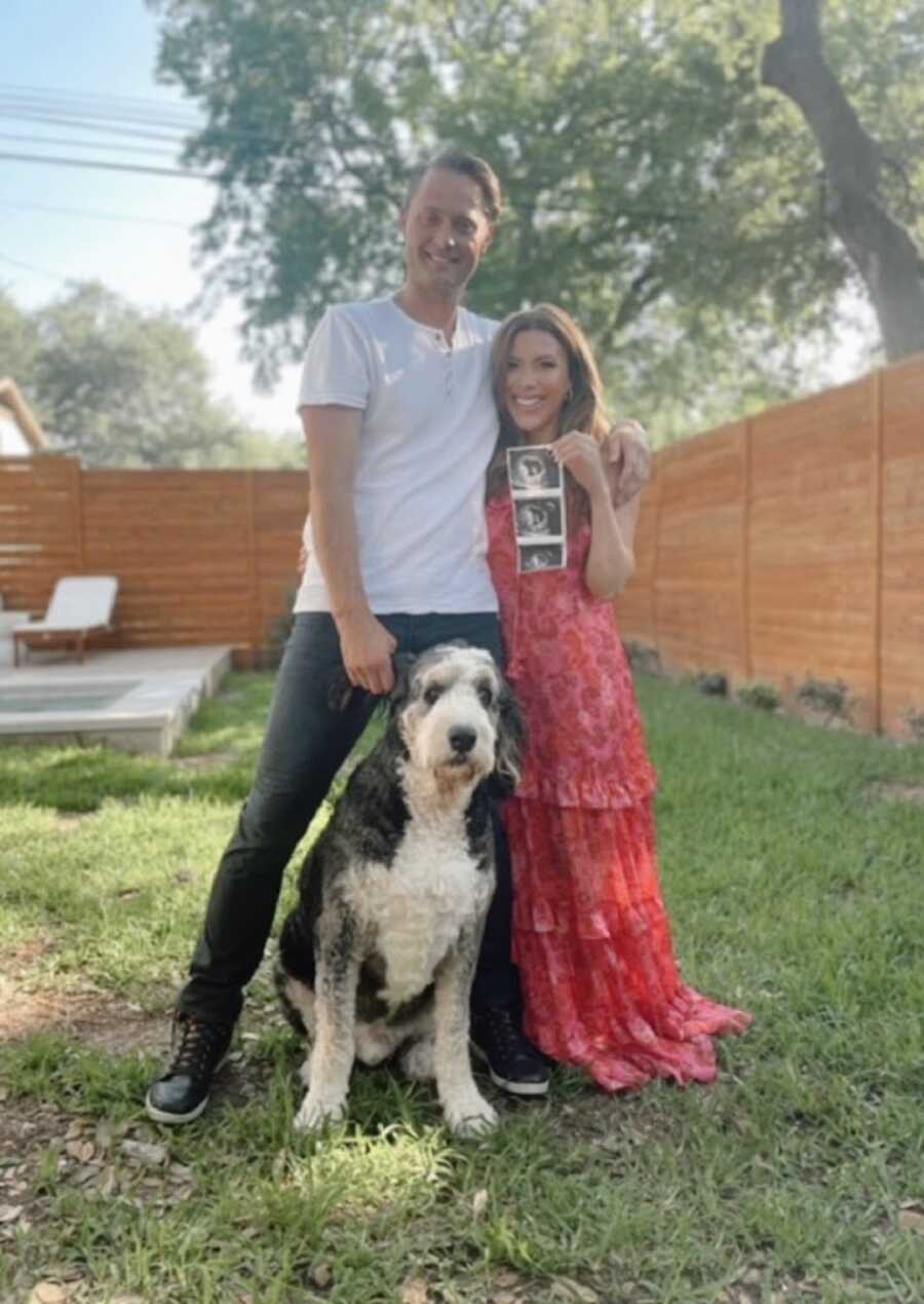 A woman, her husband, and their dog stand outside holding up sonogram pictures