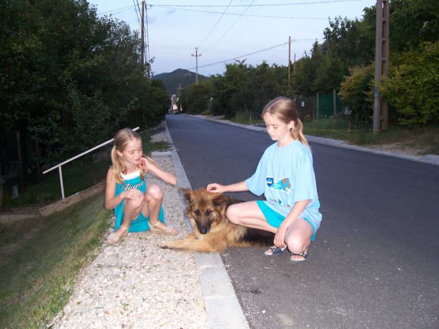 two young girls crouch at side of road and pet their dog