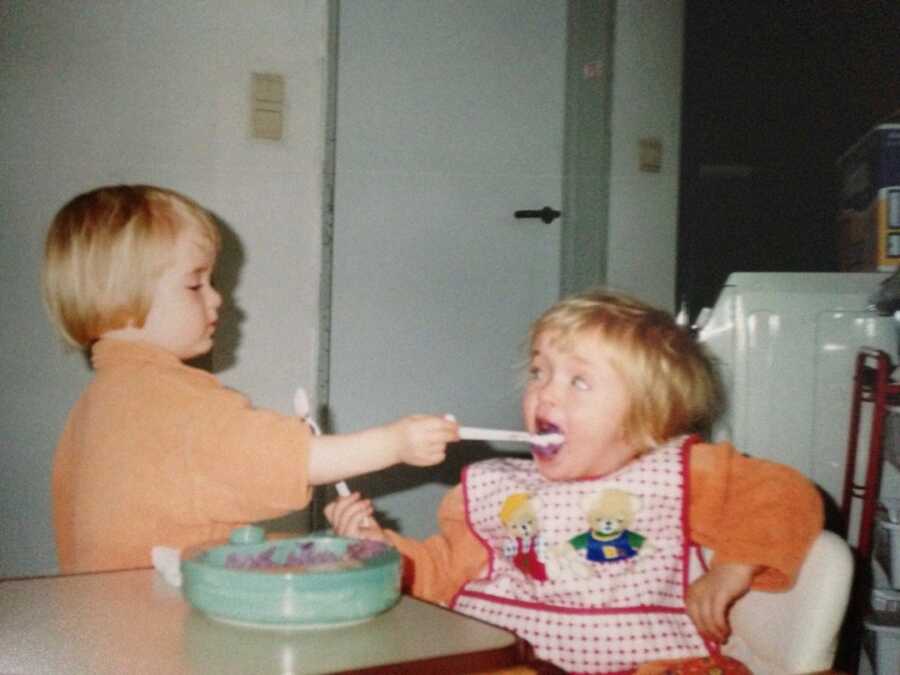 young girl sits in highchair and is being fed by a sibling