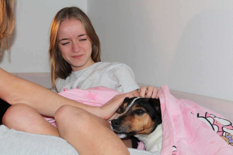girl lays in bed with her dog at her side under the covers