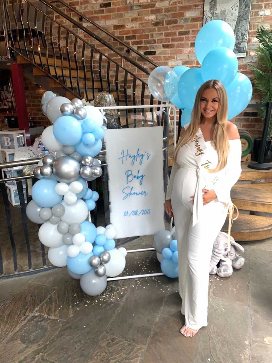 pregnant mom stands in front of sign with balloons for baby shower