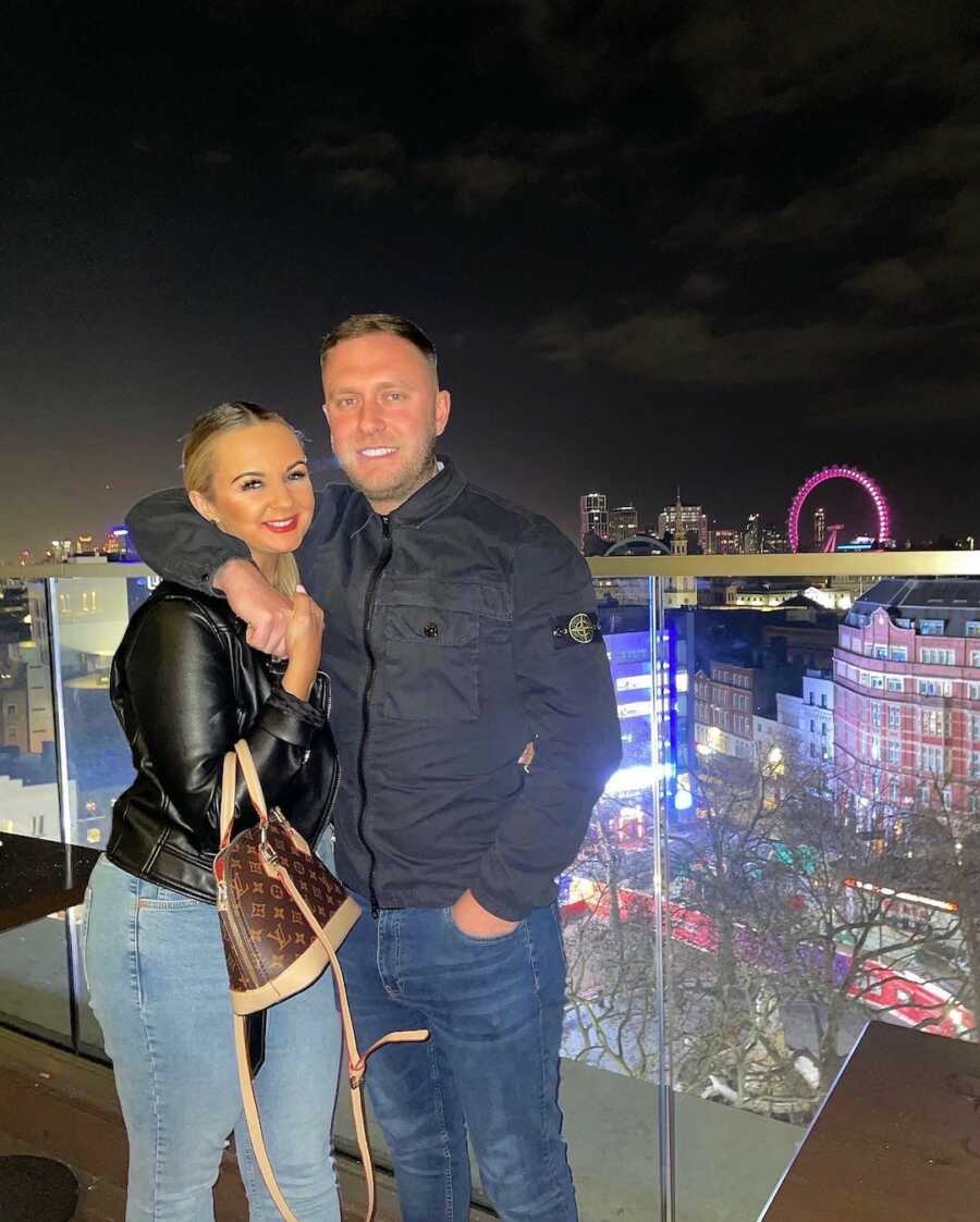 couple stands on balcony overlooking city holding one another