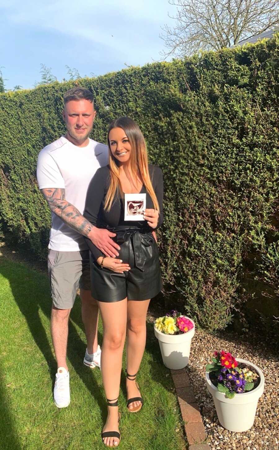 husband and wife stand holding an ultrasound image of their baby