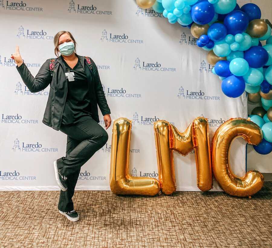 Travel nurse poses in front of the logo for a new hospital assignment