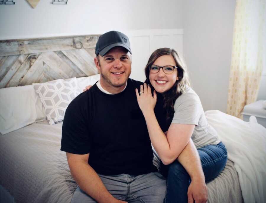 couple sits together on the corner of their bed, smiling