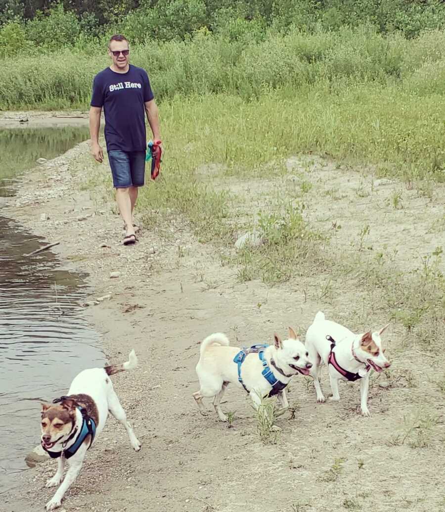 Man walks three small dogs outside along the edge of a pond.