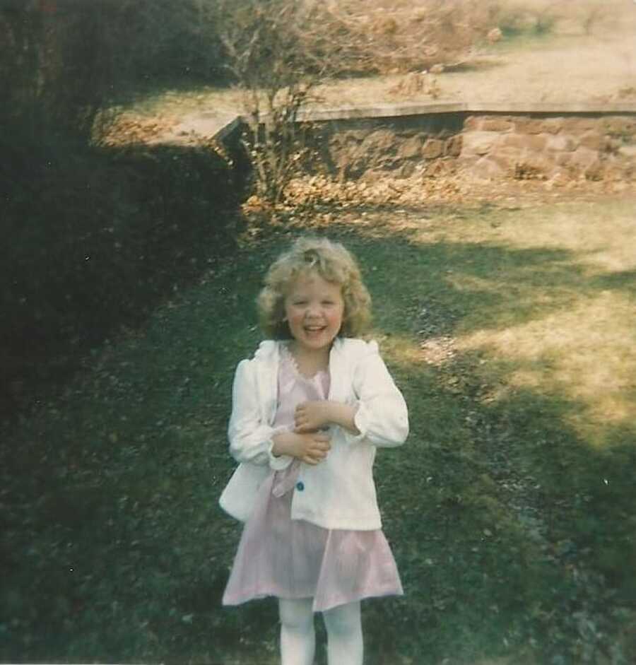 A young girl in a pink dress stands in a backyard. 