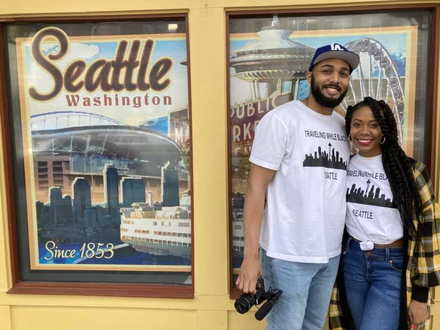 A couple in 'Traveling While Black' shirts stand in front of a Seattle poster