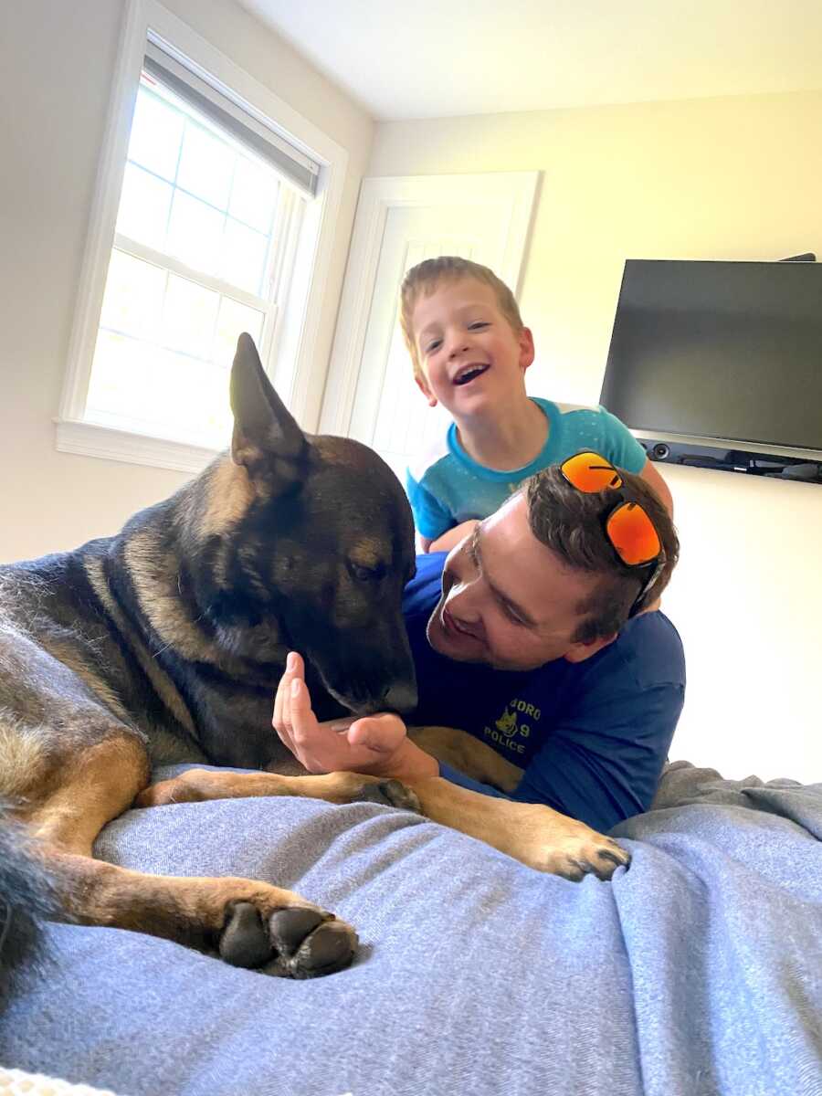 dad lays in bed with son on his back and police dog at his side