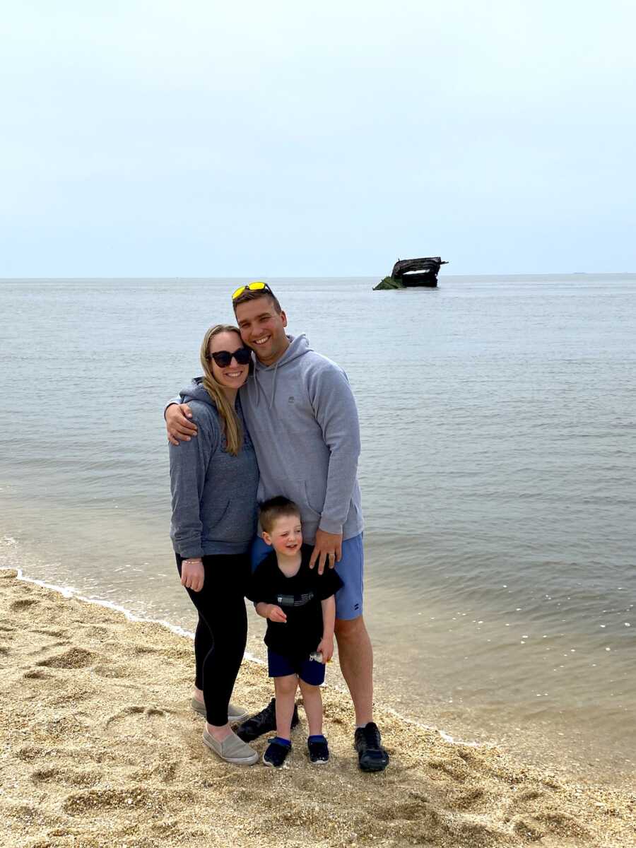 parents stand with their son in front of them, dad has his hand around wife and on son