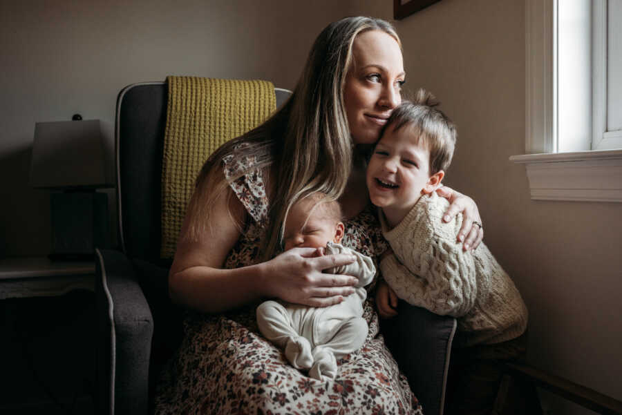 mom sits in a chair with her newborn in her lap and older son to the side of her