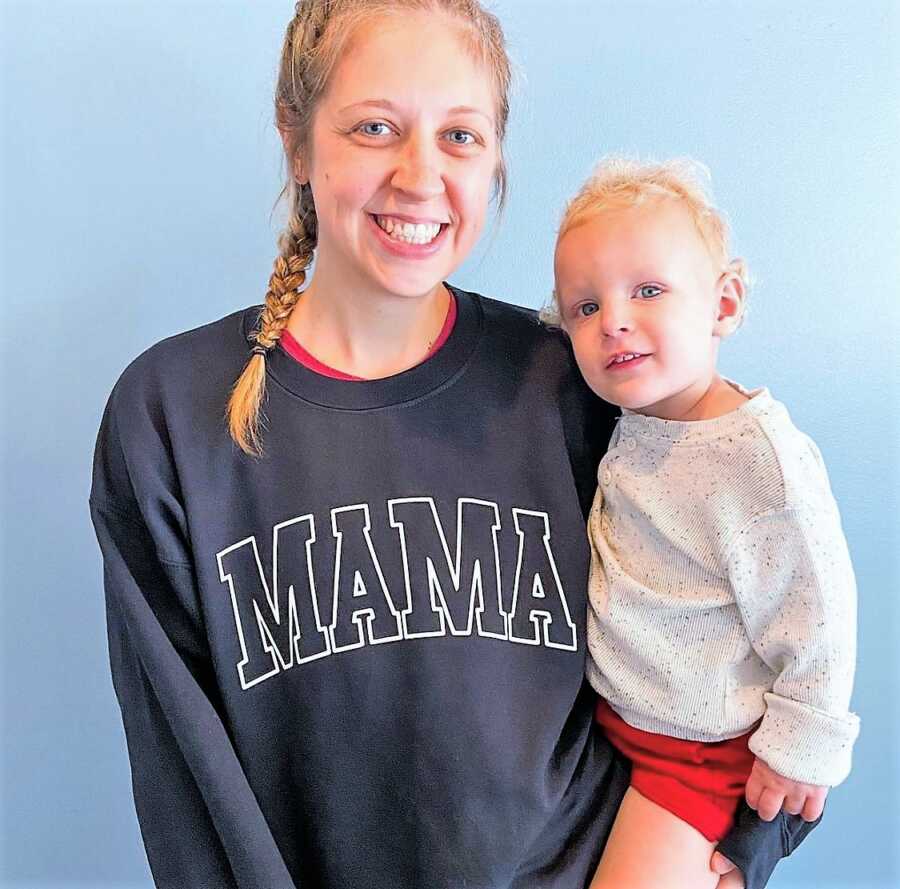 Stay-at-home mom wearing a sweatshirt that says "MAMA" while carrying her toddler son 