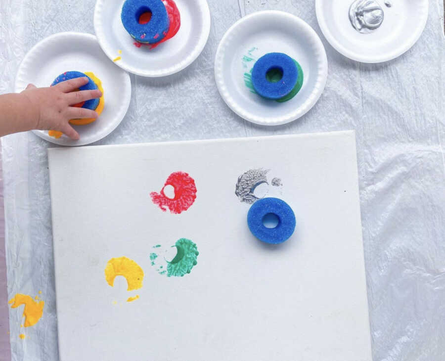 Toddler painting with a round sponge on a white canva during tot school day
