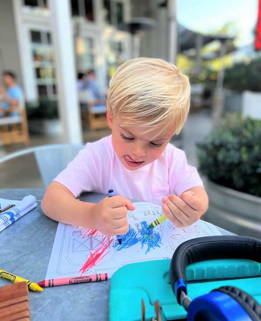 blonde, toddler boy coloring a book with crayons sitting on a patio table of a restaurant 