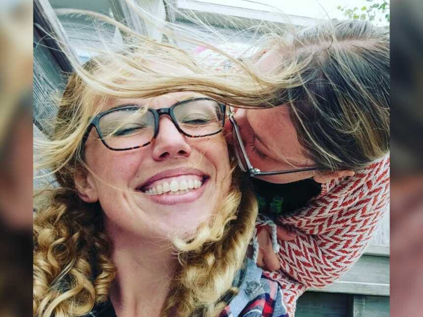Friends kisses woman on the cheek as she closes her eyes and smiles.