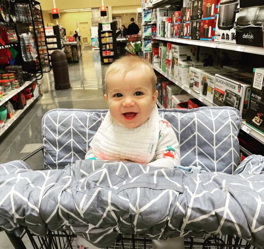 Baby sits in padded grocery cart seat.