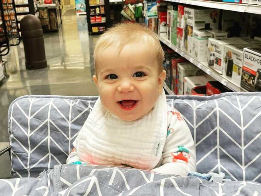 Baby sits in padded grocery cart seat.