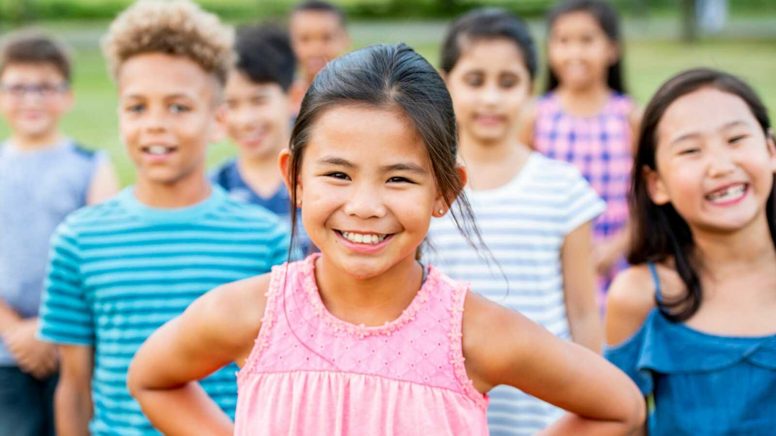 A young Asian girl stands in front of a group of diverse children with her hands on her hips and a confident smile.