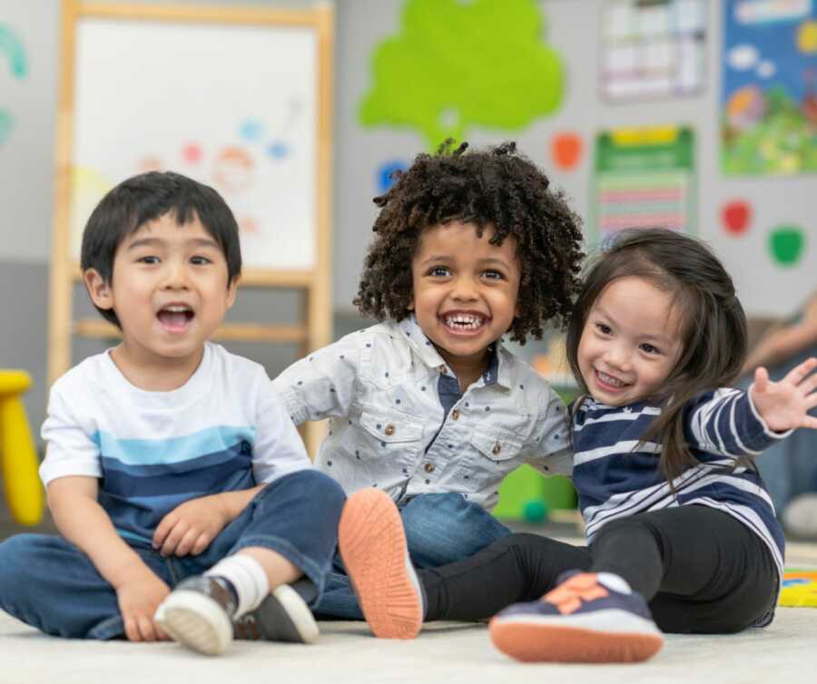 Three friends sit together laughing on the floor of their preschool.