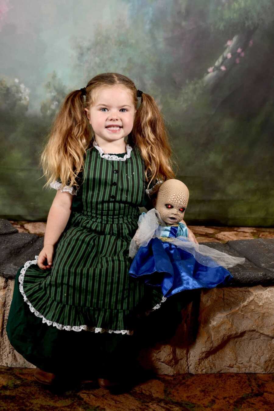 girl smiles with creepy halloween doll in dress