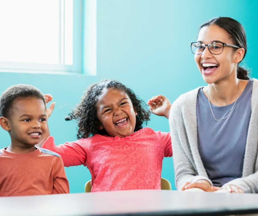 A preschool teacher and her two students smile and laugh while sitting at a table together.