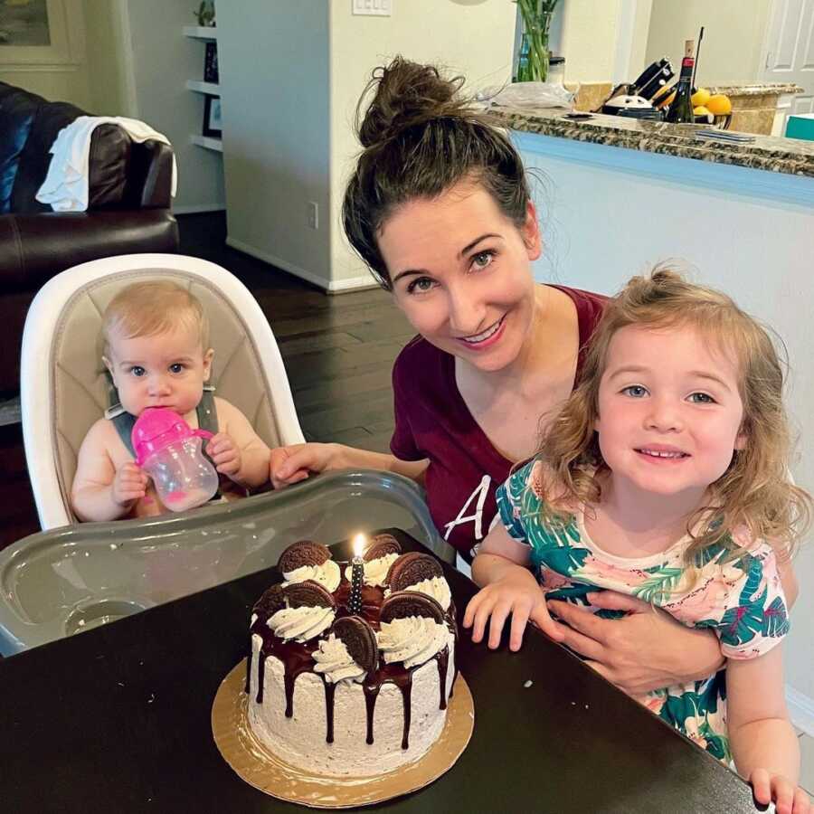 Mom holds daughter and sits next to baby in highchair for picture with birthday cake.