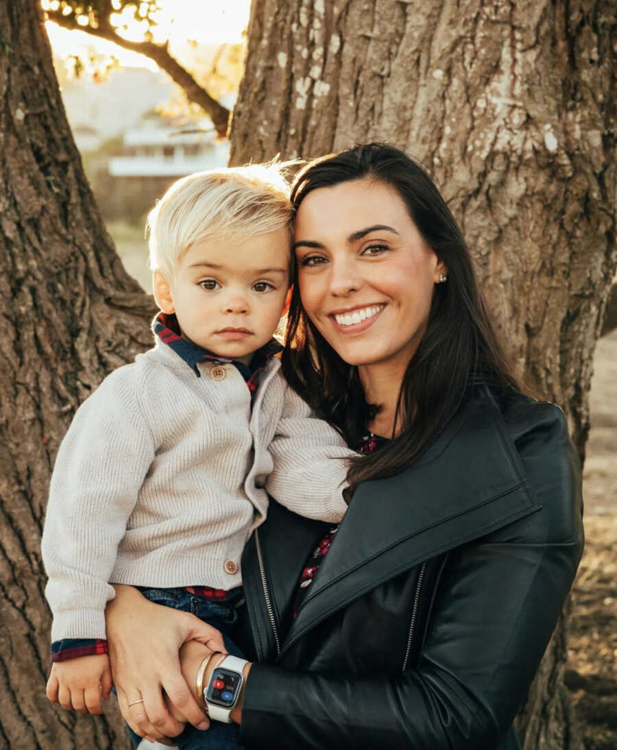 First-time mom holding toddler son in her arms in front of a large tree 