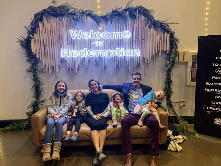 parents and their four daughters sitting on a couch behind neon sign