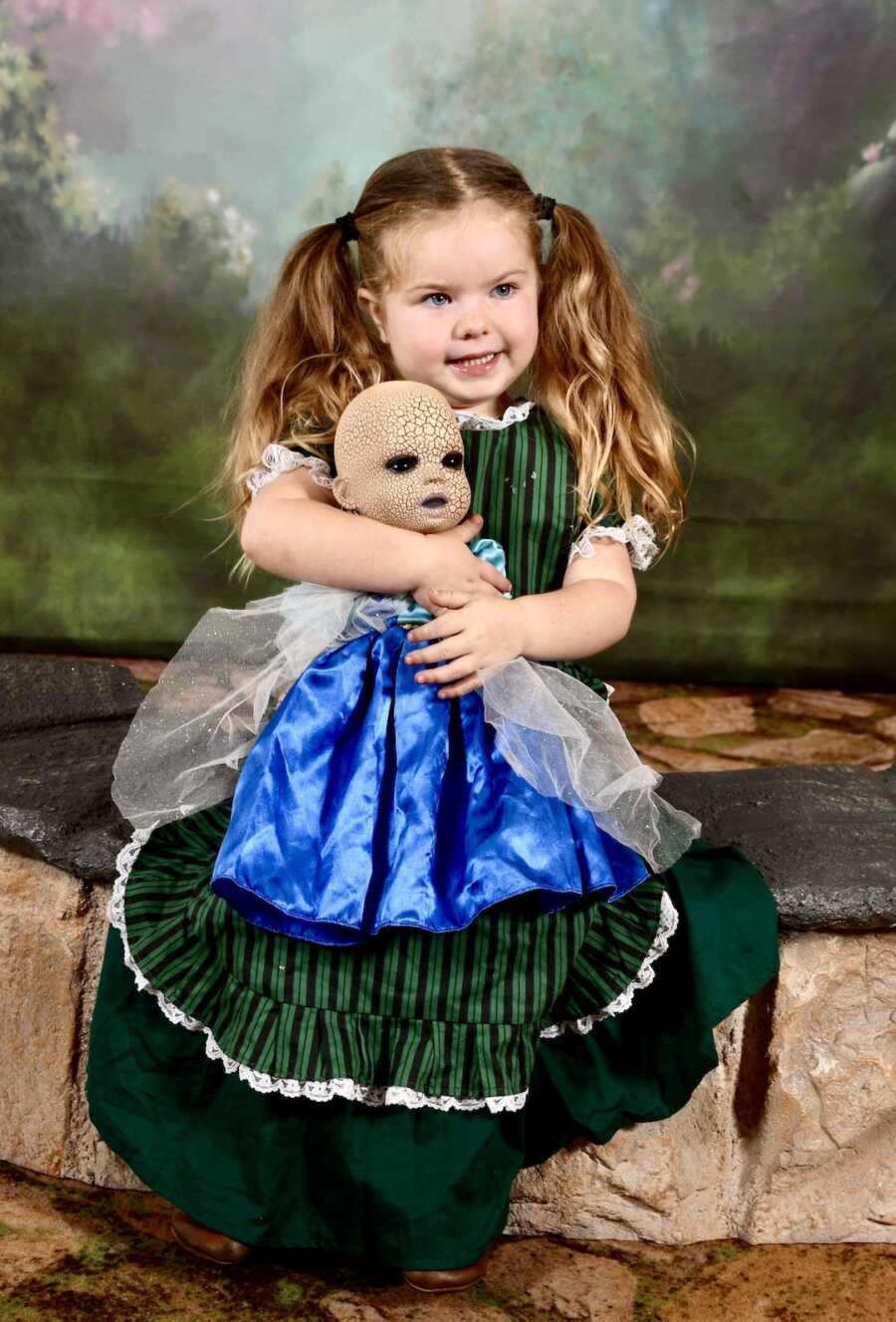 girl in green dress holds creepy doll in blue dress and smiles