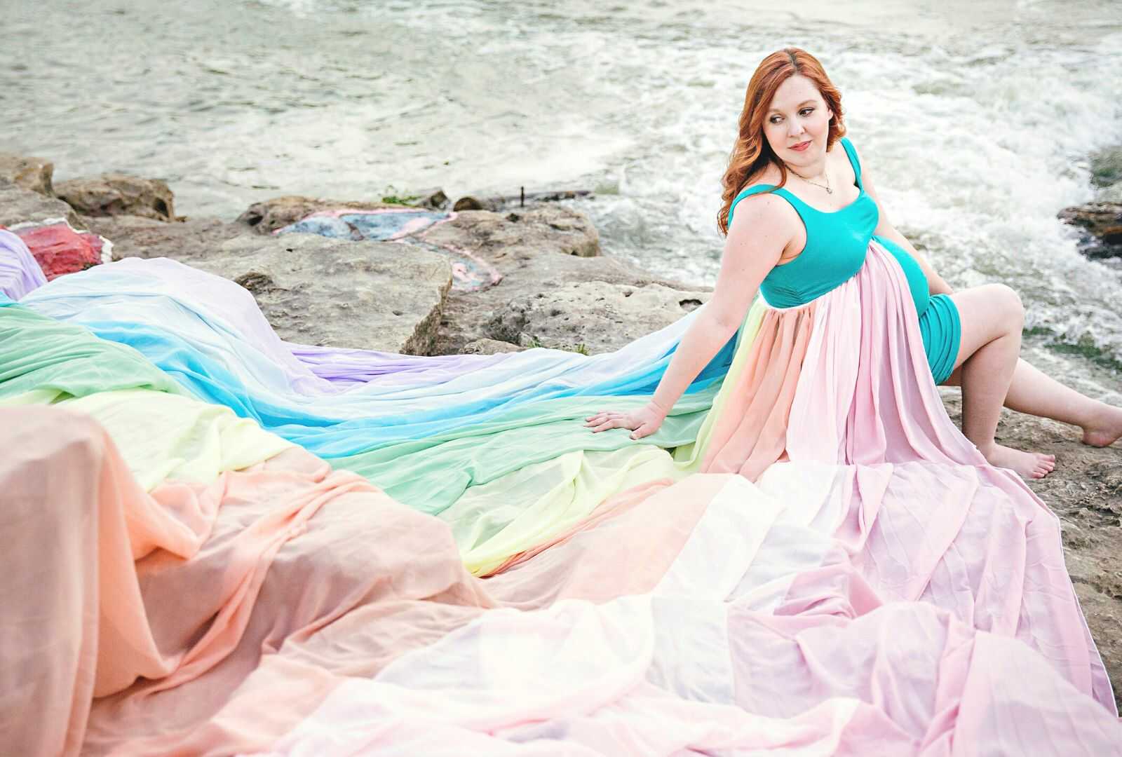pregnant mother wearing a rainbow skirt sitting on ocean rocks