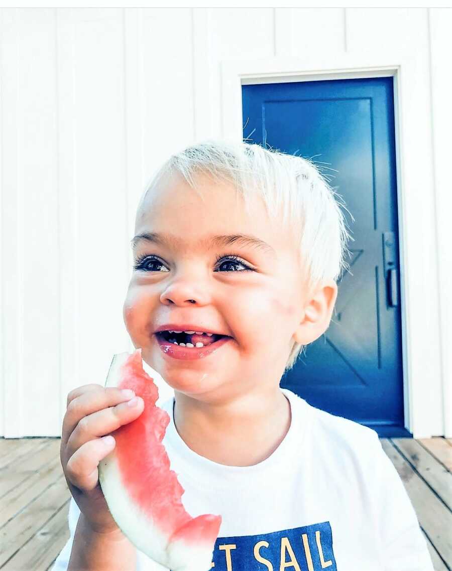 blonde, toddler boy eating a slice watermelon with a big smile on his face
