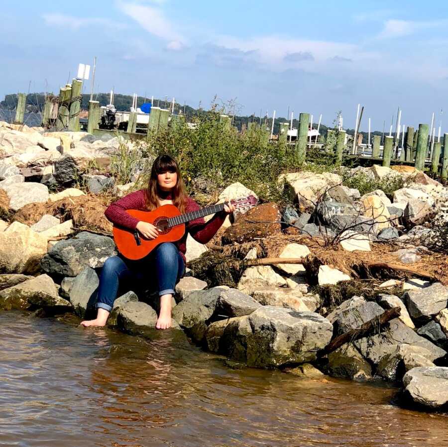 chronically ill woman sitting on rocks playing guitar