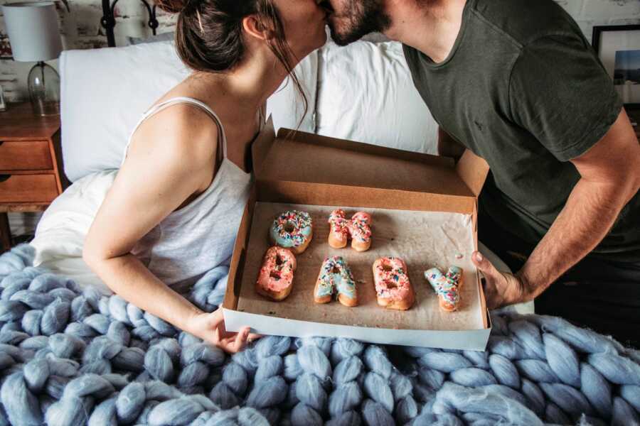 wife and husband in bed kissing over a box of donuts for pregnancy announcement