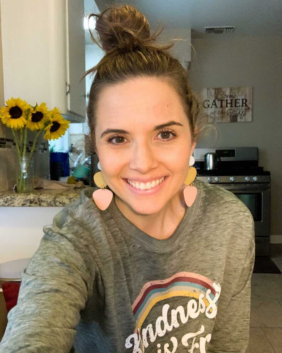 Woman wearing a kindness sweatshirt smiles for a selfie in her kitchen.