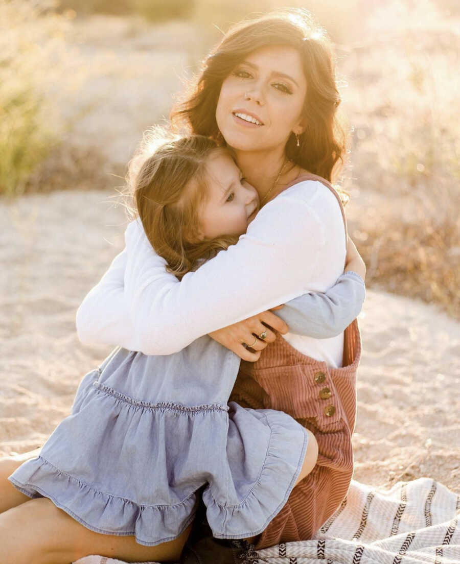 Mom hugging her young daughter outdoors with sun shining in back 