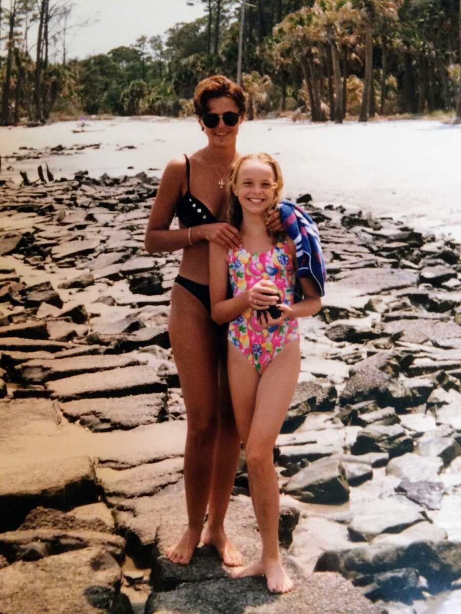 alcoholic mother and her daughter in swimsuits standing on rocky beach by a lake