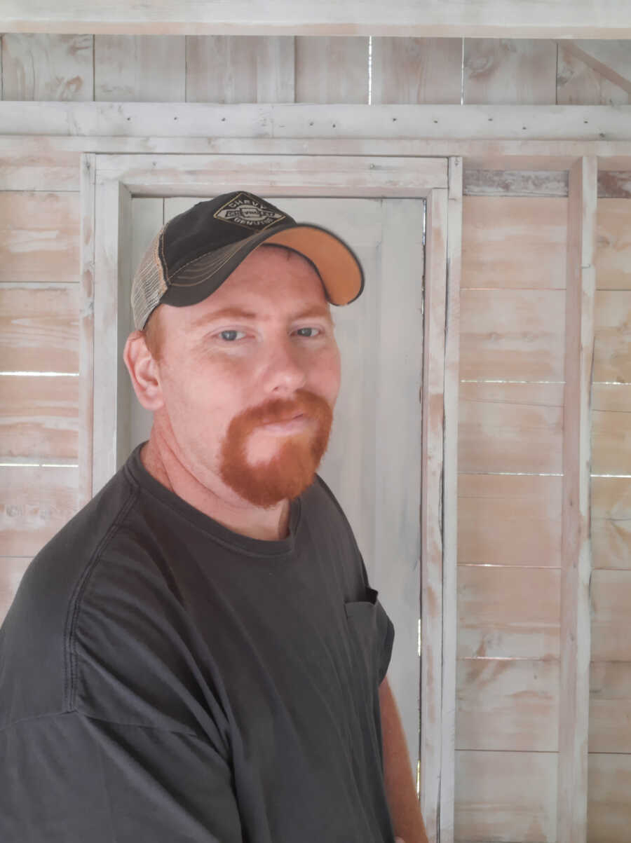 man battling melanoma stands while wearing a hat