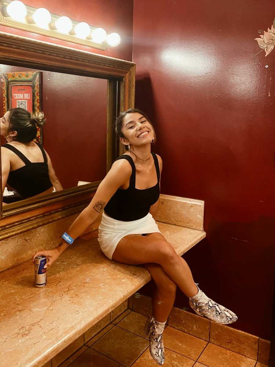 sober young woman wearing white skirt and black top sitting on bathroom counter smiling at camera