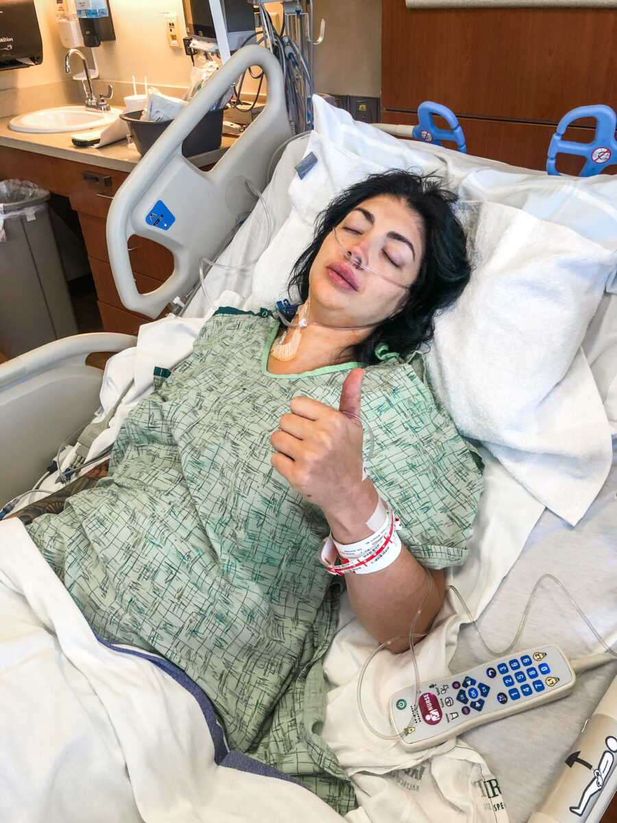 Woman in hospital bed giving thumbs up post open heart surgery
