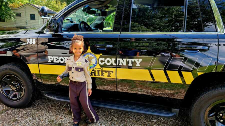 Little girl wearing a police uniform costume standing next to black sherrif truck