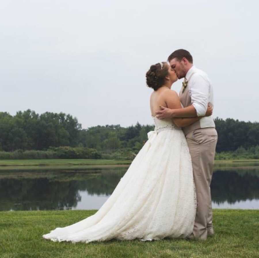 husband kisses wife in front of pond on wedding day