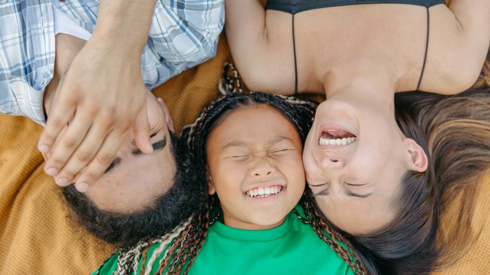 parents and child laughing while laying on yellow blanket